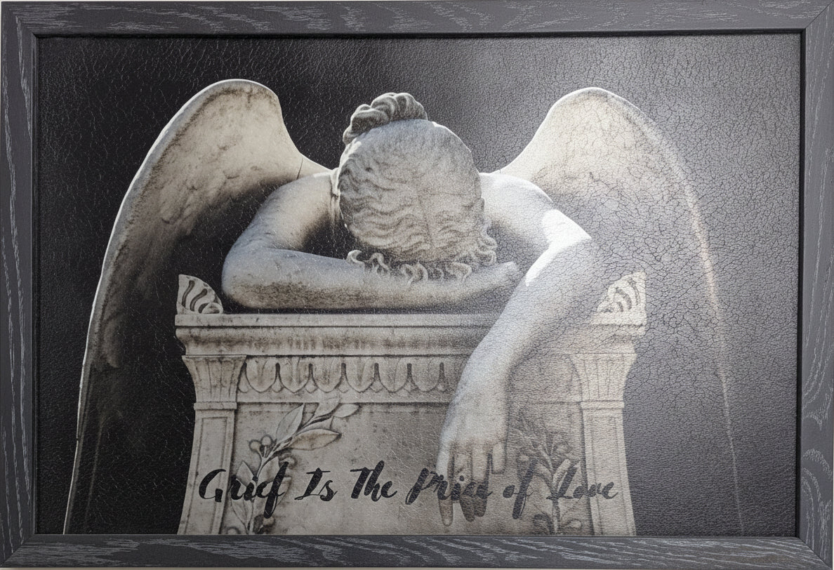 Framed artwork of an angel with wings closed, sitting on a tombstone with text.