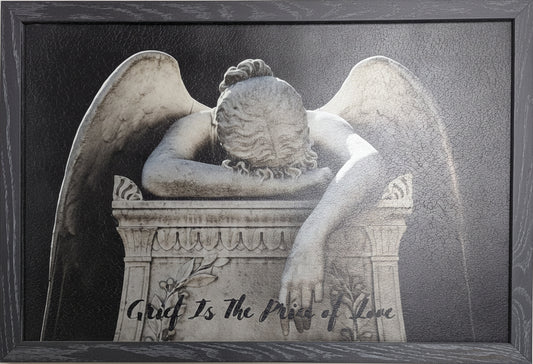 Framed artwork of an angel with wings closed, sitting on a tombstone with text.