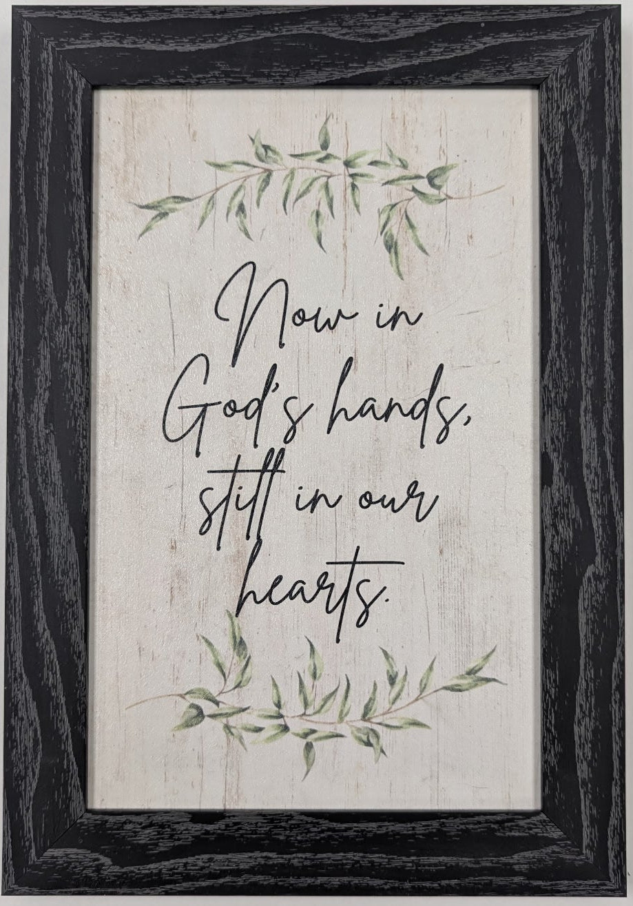 Framed print with motivational quote 'Now in God's hands, still in our hearts' on a white wall.