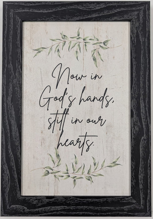 Framed print with motivational quote 'Now in God's hands, still in our hearts' on a white wall.