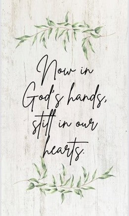 Text 'Now in God's hands, still in our hearts' with green leaves on a light wooden background